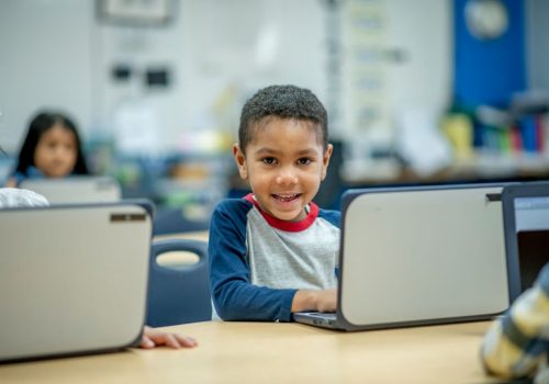 A cute African American boy using his laptop in his preschool classroom. The boy is smiling directly into the camera. In the background, there are other children also using their laptops.